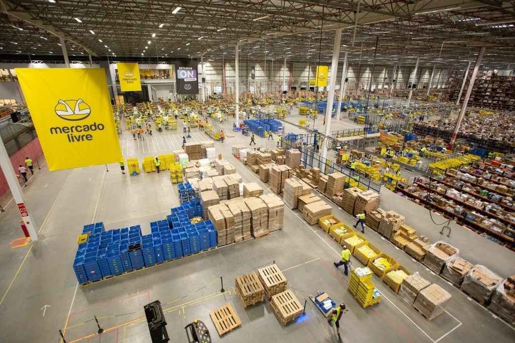 Centro de distribuição do Mercado Livre com pedidos em caixas, paletes e operadores em movimento
